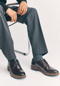 Chaussures habillées en cuir bordeaux foncé avec lacets, bout rond et semelle en cuir marron. Pantalon gris et chaussettes visibles, assis sur une chaise chromée.