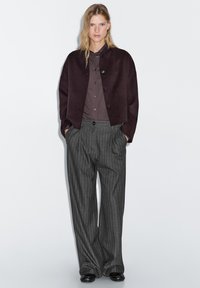 Bordeaux cropped jack over een donker overhemd met knopen, gecombineerd met grijze, fijn gestreepte hoge taille broek en zwarte schoenen.