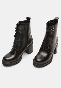 Botine negre din piele, cu toc masiv. Caracteristicile includ șireturi în față, fermoar pe lateral și talpă din cauciuc texturată pentru aderență.
