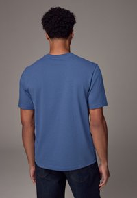 T-shirt s kratkimi rokavi in okroglim izrezom iz enobarvnega modrega bombaža. Mehka tekstura, sproščen kroj in raven rob. V kombinaciji s temnimi kavbojkami.