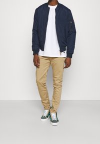 Giacca bomber navy con polsini a costine, t-shirt bianca a girocollo, chinos beige e sneakers con dettagli verdi. Tessuto morbido e vestibilità casual.