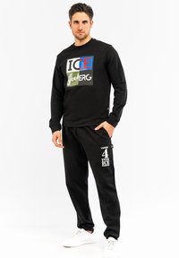 Crna dukserica s višebojnim tekstom i logotipom, u kombinaciji s crnim trenirkama koje imaju bijeli print. Obe su izrađene od mekanog materijala.