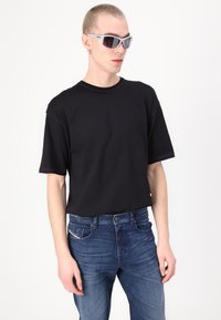 T-shirt noir à manches courtes avec des coutures contrastantes rose, associé à un jean bleu présentant un design de poche distinctif. Modèle portant des lunettes de soleil.