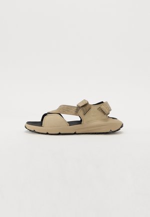 Beżowy sandał na otwartym palcu z dwoma regulowanymi paskami, czarną miękką podeszwą i wytrzymałym bieżnikiem, pokazany z boku na białym tle.