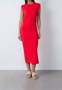 Robe midi rouge à manches courtes, design ajusté, texture lisse. Associée à des sandales tressées beiges. Fond neutre.