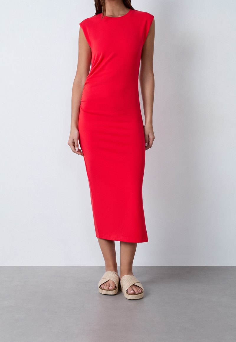 Robe midi rouge à manches courtes, design ajusté, texture lisse. Associée à des sandales tressées beiges. Fond neutre.