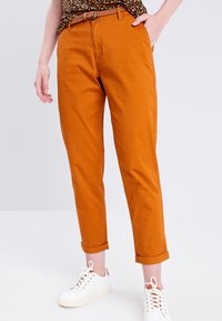 Pantalon chino orange en coton, avec une coupe décontractée, des revers roulés, porté avec un haut à motifs et des baskets blanches.