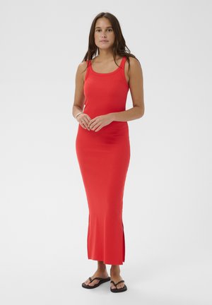 Kobieta stojąca ze złączonymi rękami, ubrana w czerwoną, bezrękawnikową sukienkę maxi i czarne klapki, na tle jasnego, jednolitego tła.