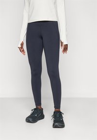 Person med mørkeblå leggings, svarte joggesko og en langermet hvit topp med tommelhull, som står mot en enkel lys bakgrunn.