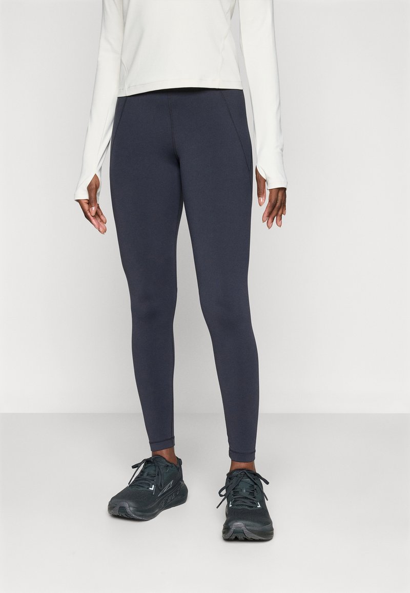 Person med mørkeblå leggings, svarte joggesko og en langermet hvit topp med tommelhull, som står mot en enkel lys bakgrunn.