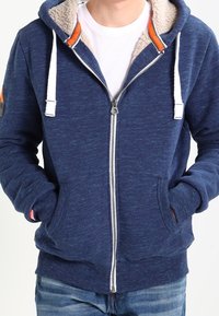 Sudadera con cremallera de color navy con cordones blancos y forro de sherpa, que presenta dos bolsillos frontales y acentos en naranja contrastante. Tejido de mezcla de algodón.