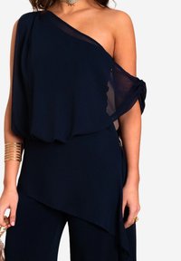 Haut asymétrique bleu marine en matériau transparent avec un design à une épaule et un tissu drapé. Présente un accent noué à la manche.