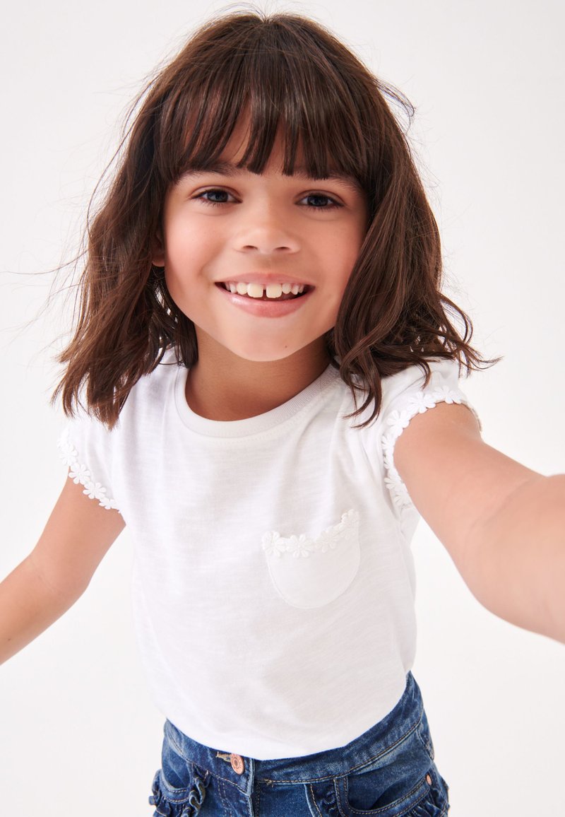 Lächelndes junges Mädchen mit braunen Haaren, das ein weißes Shirt mit floralem Rand und blaue Jeansshorts trägt und sich mit ausgestreckter Hand zur Kamera vor weißem Hintergrund reicht.
