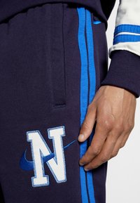 Marinblå joggare med blå sidostreck, med en stor vit broderad "N"-logotyp och Nikes karaktäristiska checkmark-ackent. Mjuk tygkänsla.