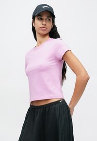 Jasnoróżowy krótki t-shirt z miękkiego materiału, z małym białym logo Nike na piersi. W zestawie z czarnymi plisowanymi spodniami.