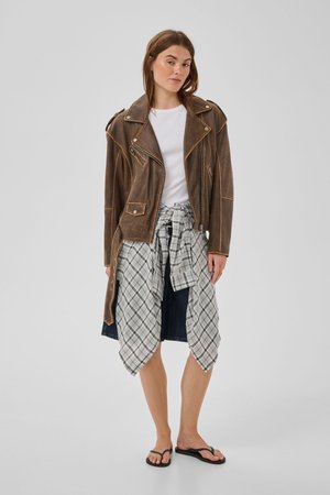 Femme portant une veste en cuir marron, un t-shirt blanc, une chemise à carreaux nouée autour de la taille, une jupe bleu marine et des tongs noires, debout devant un fond uni.