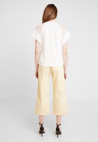Camiseta blanca con panel de encaje en los hombros, combinada con pantalones de pierna ancha de un color amarillo claro y sandalias de tacón negras. Diseño simple y relajado.