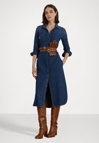 Abito camicia in denim con chiusura anteriore a bottoni, maniche lunghe e una cintura in pelle marrone dotata di portaguanti. Stivali con tacco marroni.