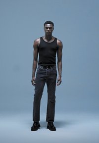 Camiseta sin mangas negra, jeans de mezclilla oscura, botas negras; modelo de pie contra un fondo azul claro sólido, mostrando un diseño minimalista.
