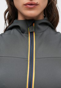 Casaco desportivo cinzento com capuz, com um fecho éclair multicolorido riscado em vermelho, amarelo e azul. Textura lisa, design ajustado.