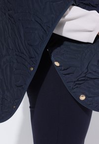 Gesteppte marineblaue Jacke mit floraler Steppung und goldenen Druckknöpfen, getragen über einem weißen Pullover und marineblauen Hosen.