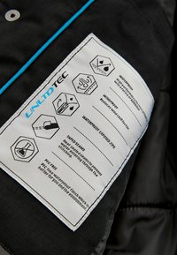 Etichetta di giacca in tessuto nero con testo bianco e blu. I simboli indicano proprietà impermeabili, antivento e senza PVC. Sezioni nastrate indicate.