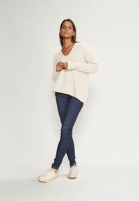 Beige gebreide trui met een diepe V-hals, losse pasvorm, lange mouwen, gecombineerd met donkerblauwe skinny jeans en lichtroze sneakers.