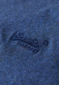 Navy blauwe katoenen t-shirt met een reliëflogo op de voorkant. De stof heeft een subtiele textuur en een licht gemêleerd uiterlijk.