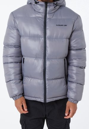 Man wearing a gray puffer jacket with a hood and black zipper, logo "CLOSURE LDN" on chest, black cargo pants visible from waist down.