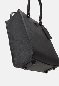 Czarna torebka o strukturze z fakturowanym wykończeniem, wyposażona w zaokrąglone uchwyty, metalowe elementy dekoracyjne oraz ochronne nóżki u podstawy.