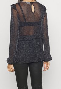 Blusa nera trasparente con luccichio argento, dettagli arricciati sulle spalle, apertura a chiave sul retro e maniche lunghe, abbinata a pantaloni di pelle neri.