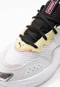 Weiße Mesh-Sneaker mit einem gepolsterten Obermaterial aus gelbem Wildleder, schwarzen Schnürsenkeln, metallischen Akzenten und pinkem Branding auf der Zunge. Verfügt über Perforationen.