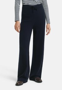 Vrouw in marineblauwe wijde broek met koord taille, gestreept lang mouwshirt en donkerbruine schoenen, staand op een witte achtergrond.
