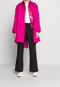 Roze waterdichte jas, oversized ontwerp, met ritszakken, gecombineerd met zwarte uitlopende broek en witte sneakers. Het kleurcontrast is duidelijk aanwezig.