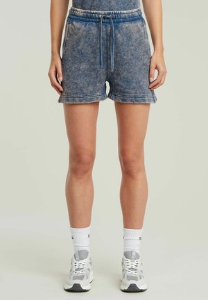 Person trägt blaue Shorts im Denim-Stil mit Kordelzug, weiße Socken und weiße Sportschuhe, steht vor einem schlichten Hintergrund.