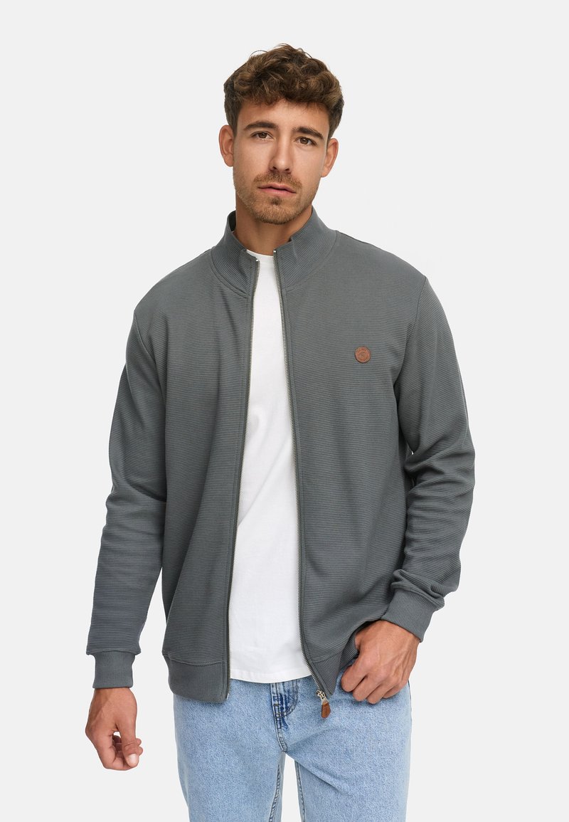 Joven con cabello corto y rizado, vistiendo una chaqueta gris con cremallera sobre una camiseta blanca y jeans de color azul claro, de pie contra un fondo blanco.