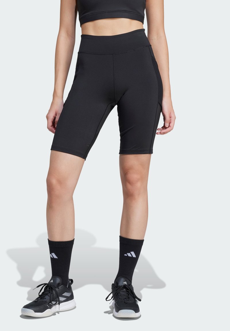 Shorts de sport noirs en tissu extensible, avec une taille haute, une texture lisse et des poches latérales. Associés à des chaussettes de sport noires et des chaussures.