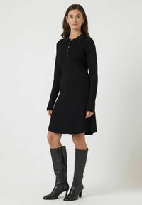 Abito lungo a maniche lunghe nero a coste con colletto, patta con bottoni e orlo leggermente svasato, abbinato a stivali alti fino al ginocchio in pelle nera.