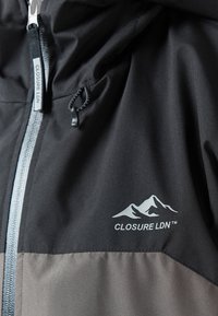 Crna i siva jakna s zatvaračem, koja ima kapuljaču na vezanje i bijelog loga s grafikom planina na prednjem dijelu.