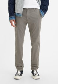 Man som bär grå byxor, svartvita sneakers, vit skjorta och en marinblå och grå vadderad jacka med denimärmar, står med händerna i fickorna.