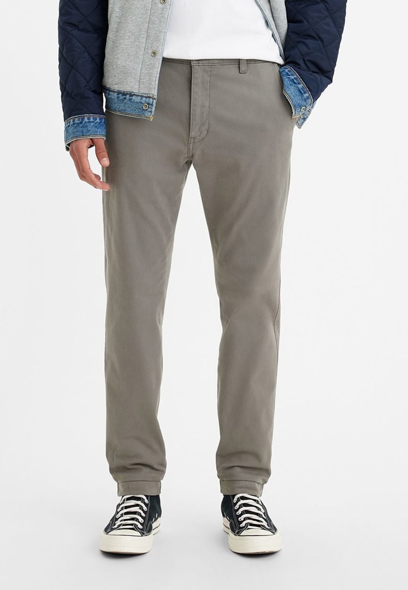 Man som bär grå byxor, svartvita sneakers, vit skjorta och en marinblå och grå vadderad jacka med denimärmar, står med händerna i fickorna.