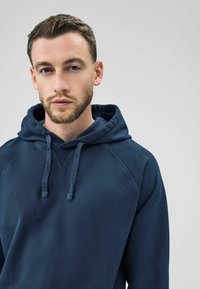 Felpa blu navy realizzata in morbido tessuto, con cappuccio regolabile, maniche raglan e vestibilità rilassata. Texture liscia con dettagli minimi.