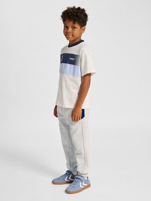 Dreng med krøllet hår iført en hvid og blåstribet t-shirt, lysegrå joggingbukser og blå sneakers, som står og smiler.