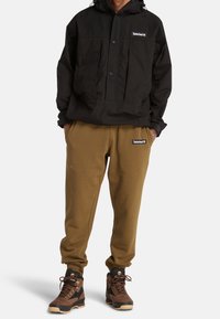 Svart vattentät jacka, bruna fleecebyxor och bruna vandringskängor med tan detaljer. Timberland-logotyper på jackan och byxorna.