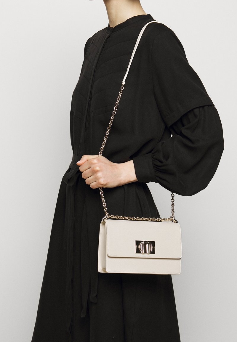Krémová kabelka s předním klopovým zavíráním a kovovým zavíráním, zdobená řetízkem. Nositá přes černý oděv s nabíranými rukávy.
