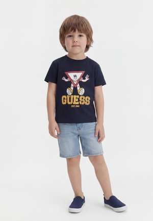 Junger Junge steht und trägt ein marineblaues T-Shirt mit einem Cartoon-Dreieck, helle Jeans-Shorts und marineblaue Slipper vor weißem Hintergrund.