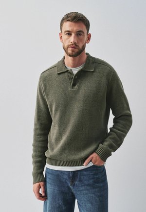 Man met kort bruin haar en baard draagt een groen gebreide trui met knoopsluiting bij de kraag over een wit overhemd en blauwe spijkerbroek, staand tegen een effen achtergrond.