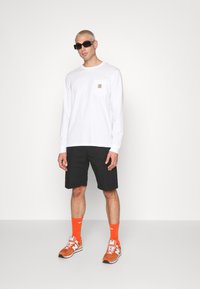 Hvit langermet skjorte med brystlomme, svarte cargo-shorts, oransje sportsokker og oransje sneakers. Modellen har på seg svarte solbriller.
