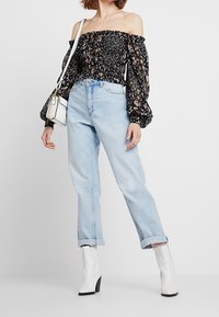 Black off-the-shoulder blouse with floral pattern, long puffed sleeves; light blue high-waisted jeans; white ankle boots with a block heel.