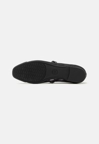 Semelle plate noire de chaussure avec prise avant texturée, talon lisse et empreinte circulaire du logo dans la zone de la voûte plantaire sur fond blanc.